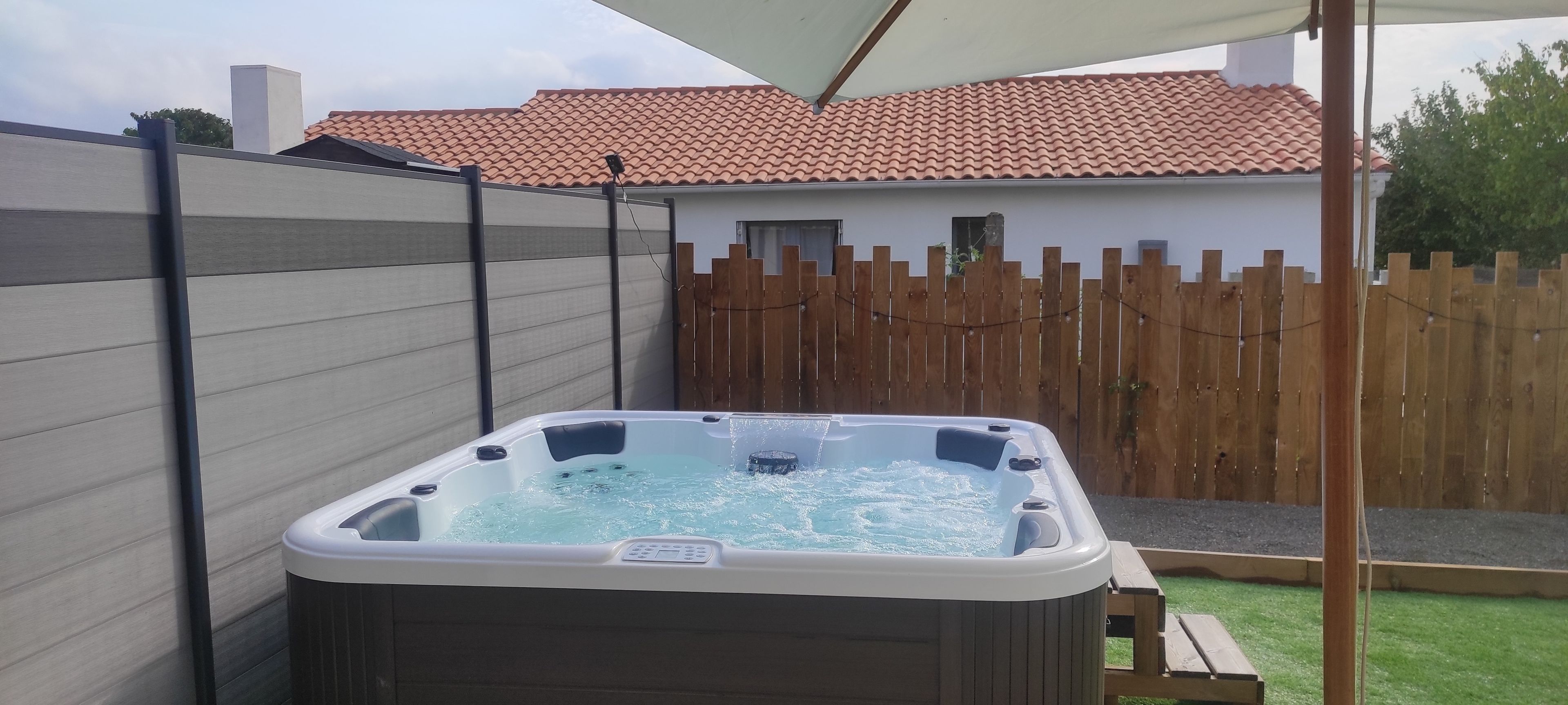 A hot tub in a backyard with a wooden fence, a white house, and a tiled roof.