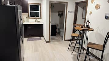 Une cuisine compacte avec des meubles de rangement sombres, un réfrigérateur et une fenêtre. On y trouve un petit coin repas avec une table et des chaises.