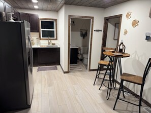 Eine kleine Küche mit dunklen Schränken, einem Kühlschrank und einem Fenster. Es gibt einen Essbereich mit Tisch und Stühlen.