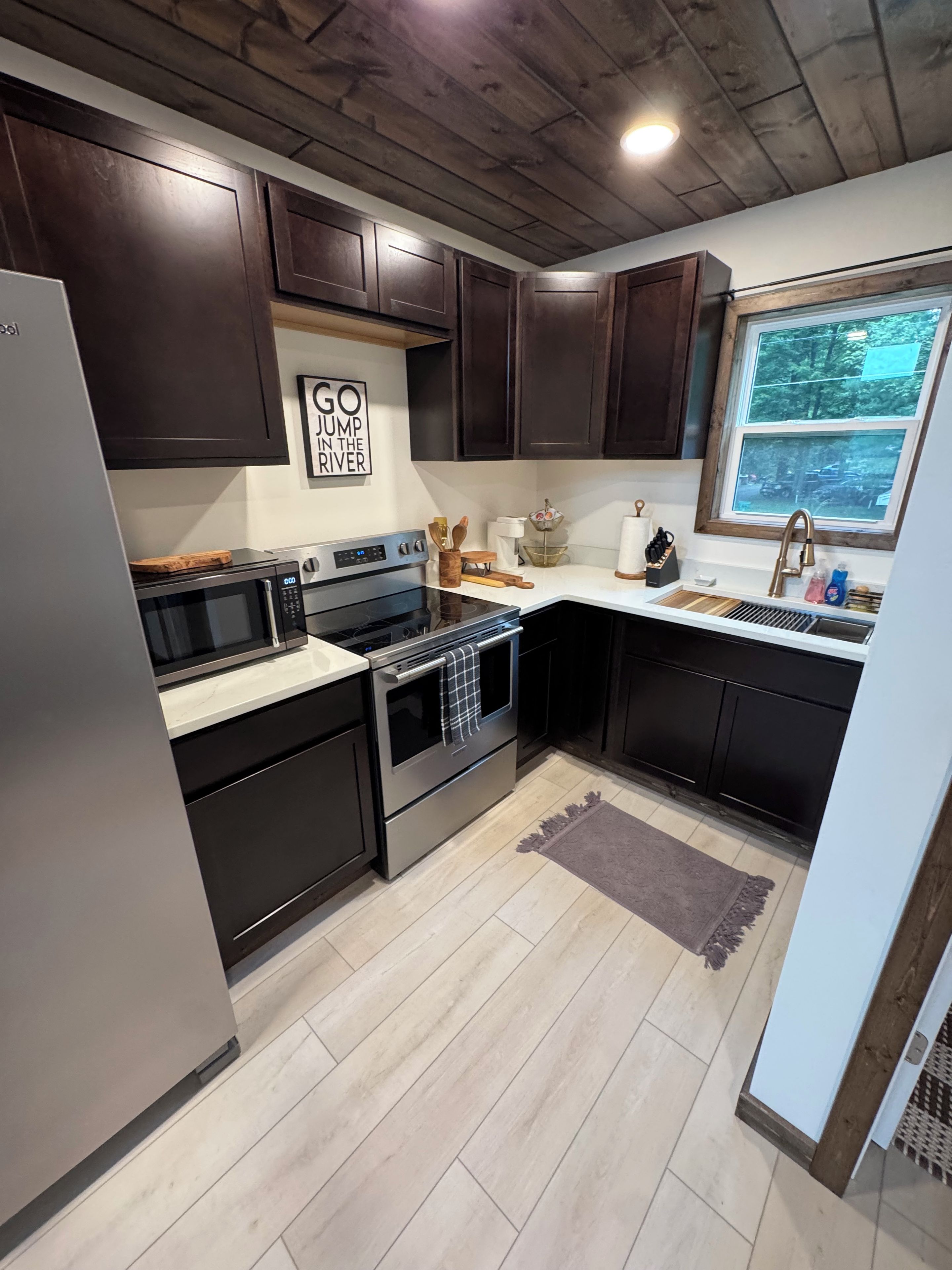 A kitchen with dark cabinets, a stainless steel refrigerator, a microwave, an oven, and a window providing a view of the outdoors.