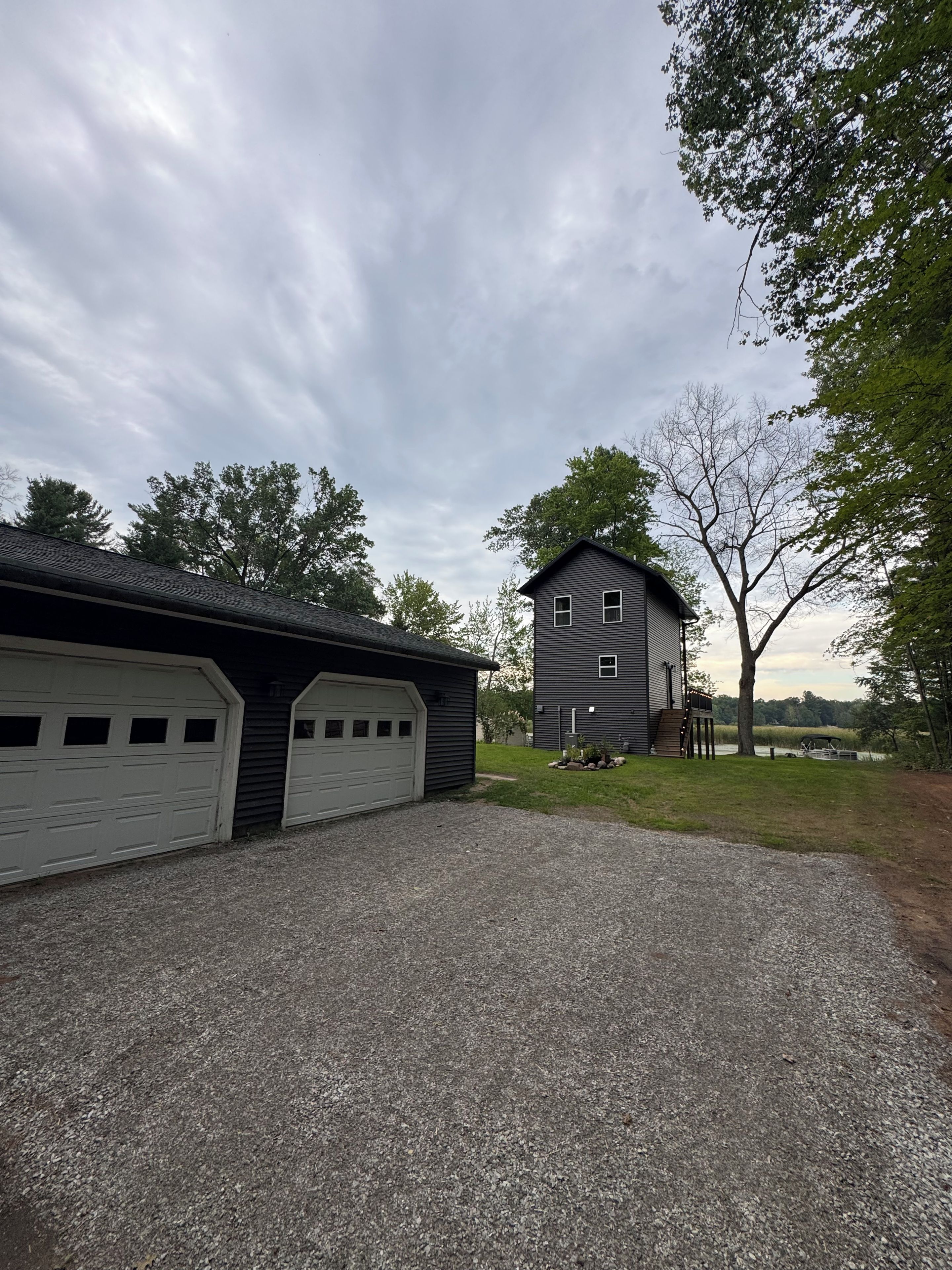 A two-story building with a garage and a covered area, surrounded by trees and a gravel driveway.