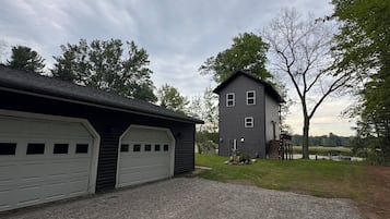 A two-story building with a garage and a covered area, surrounded by trees and a gravel driveway.