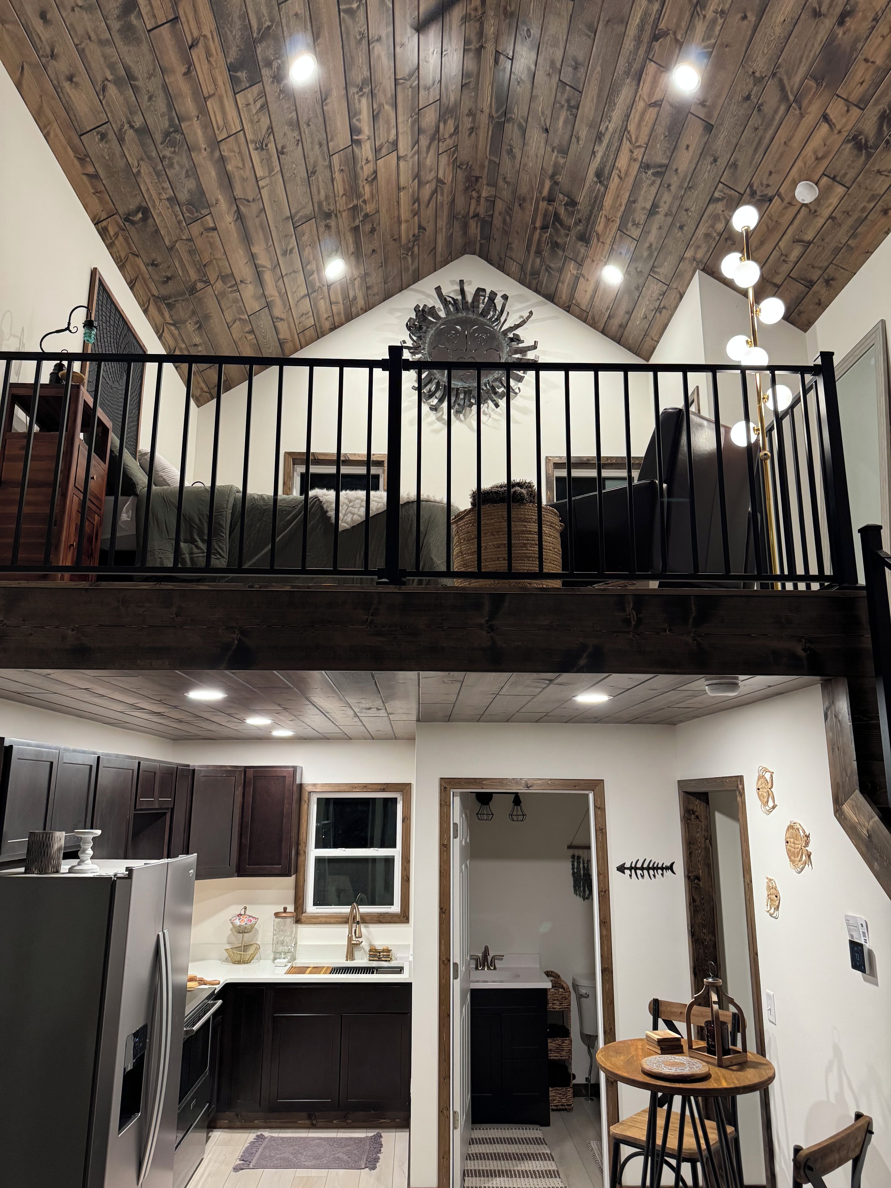 Quarto estilo loft com teto de madeira, varanda com guarda-corrimão preto e área de jantar com mesa redonda.