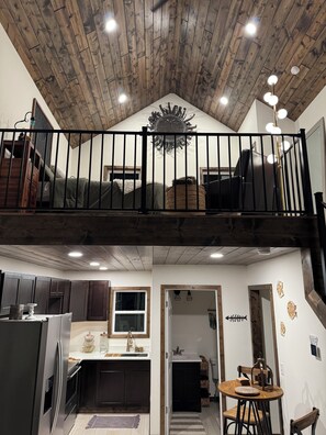A loft-style room with a wooden ceiling, a balcony with a black railing, and a dining area with a round table.