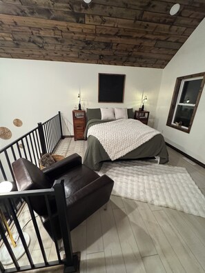 Ein Schlafzimmer mit Holzboden, einem Bett mit weißer Decke, einem Nachttisch mit Lampe, einem Fenster mit Blick und einer Treppe.