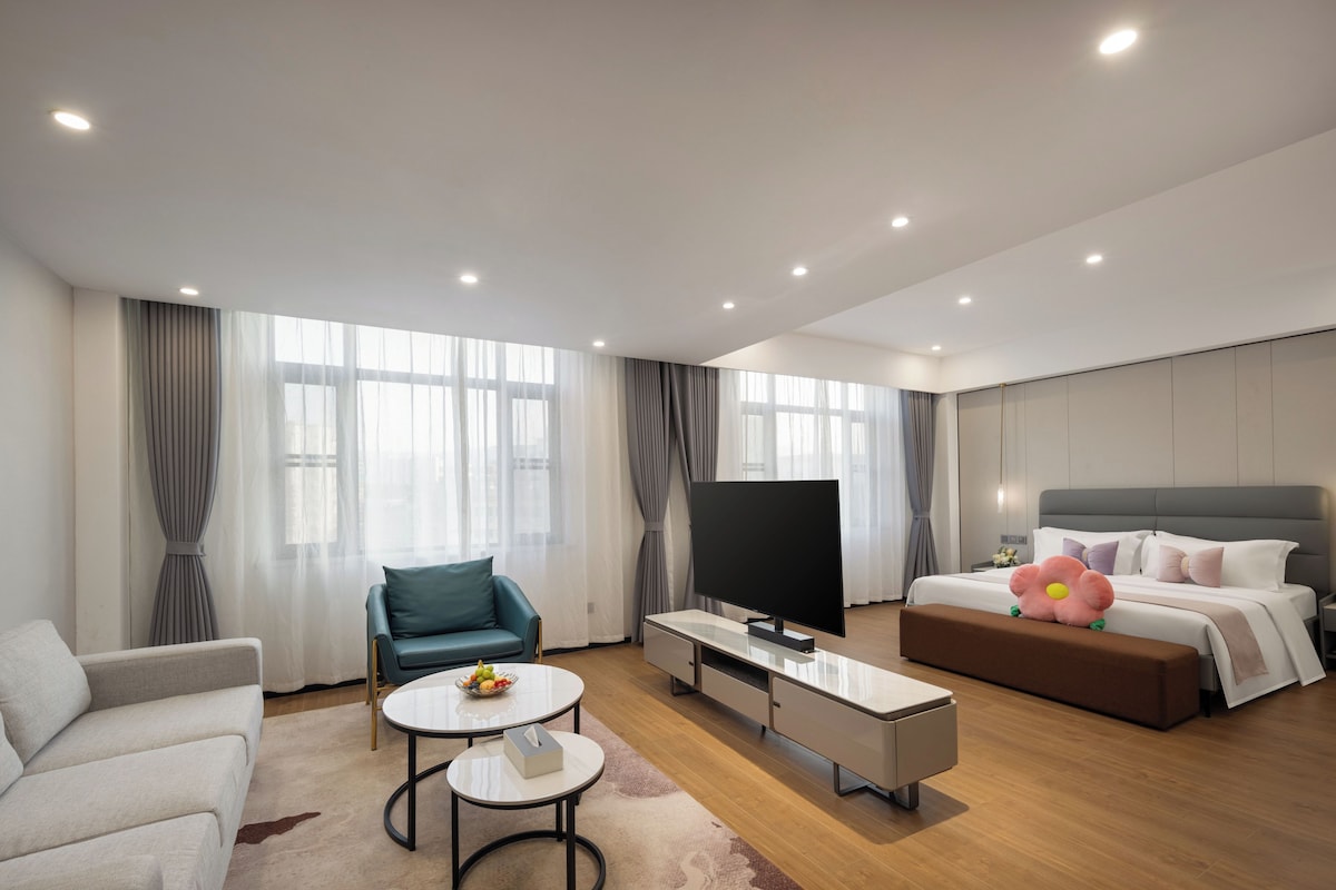 Una habitación de hotel moderna con sofá, mesa de comedor, televisión, cama y un amplio ventanal.