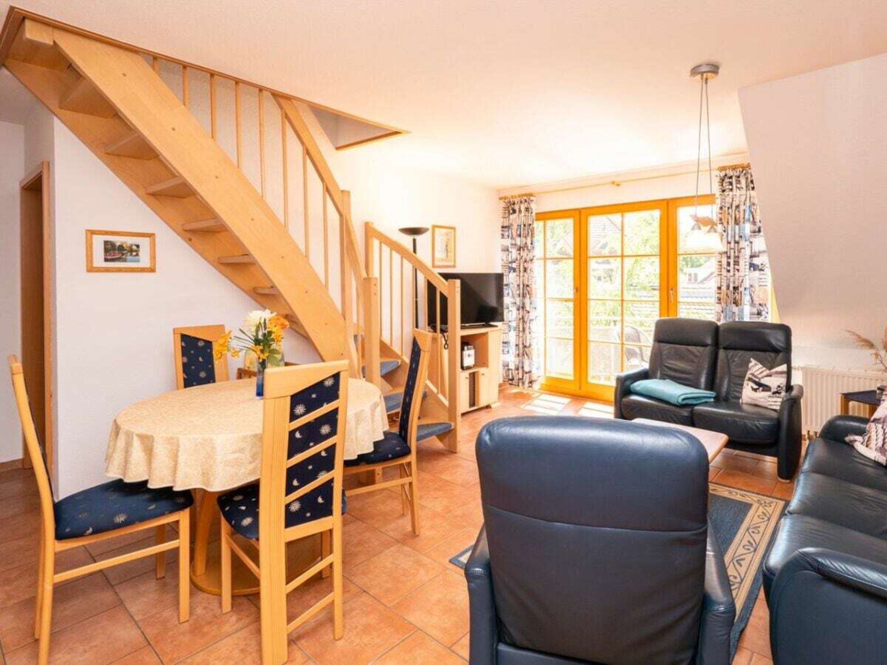 A living room with a wooden staircase, a dining area with a table and chairs, a black leather sofa, and a television.