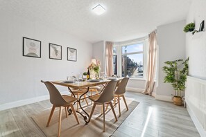 A dining area with a wooden table, four chairs, a vase with flowers, and framed artwork on the wall. - House Sleeps 7 With Enclosed Garden (Coventry)