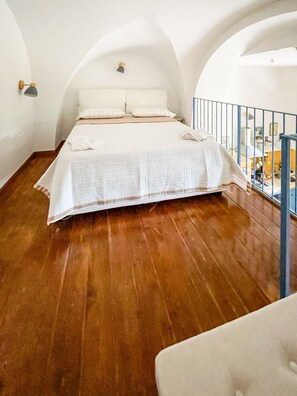 A bedroom with a wooden floor, a bed with a white bedspread, and a balcony with a railing. - Pierina's Almond Tree (Vitulano)