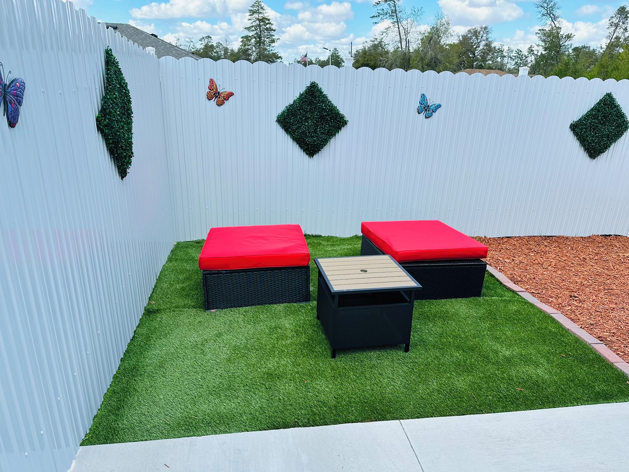 A patio area with artificial grass, two red cushioned benches, a small table, and decorative butterfly cutouts on a white fence.