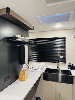 A modern kitchen with a white countertop, black cabinetry, a black backsplash, and a window with blinds.