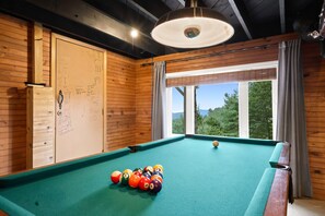 Ein Billardtisch mit Kugeln, eine Holzwand mit einer Whiteboard-Tafel und ein Fenster mit Blick auf Bäume.