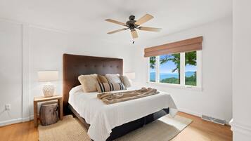 A bedroom with a bed, a ceiling fan, a window with a view, and a nightstand with a lamp.