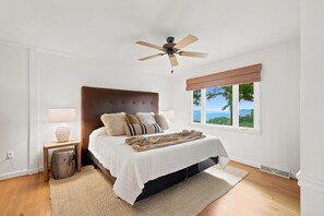 A bedroom with a bed, a ceiling fan, a window with a view, and a nightstand with a lamp.