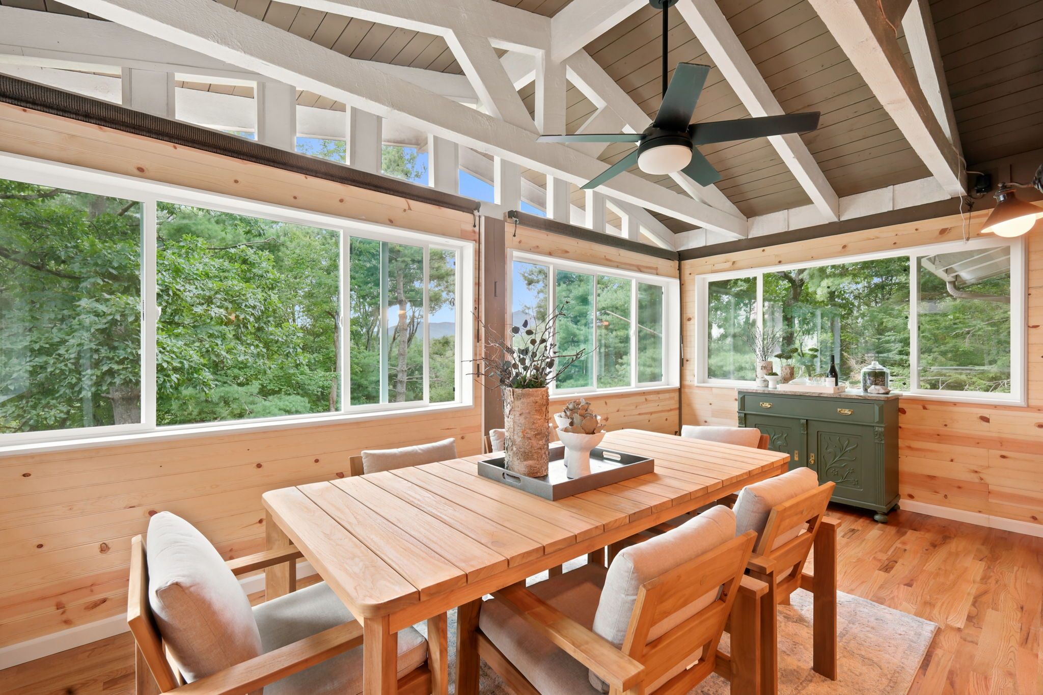 A spacious dining area with a wooden table and chairs, large windows offering a view of the outdoors, and a ceiling fan.