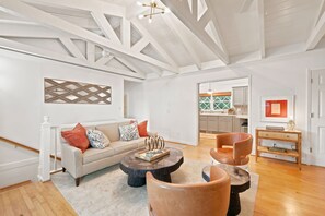 A modern living room with a sofa, coffee table, and chairs. There is a kitchen visible through an open doorway.