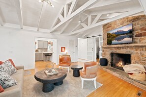 Amplia sala de estar con chimenea, piso de madera y una cocina al fondo.