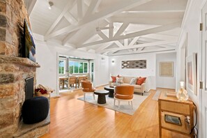 A spacious living room with a stone fireplace, wooden flooring, and a high ceiling with exposed beams.