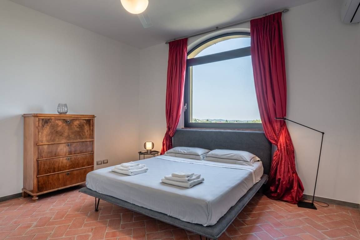 Una camera da letto con un letto, una cassettiera in legno, un comodino con una lampada e una grande finestra con tende rosse.