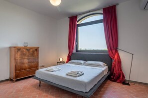 Una camera da letto con un letto, una cassettiera in legno, un comodino con una lampada e una grande finestra con tende rosse.