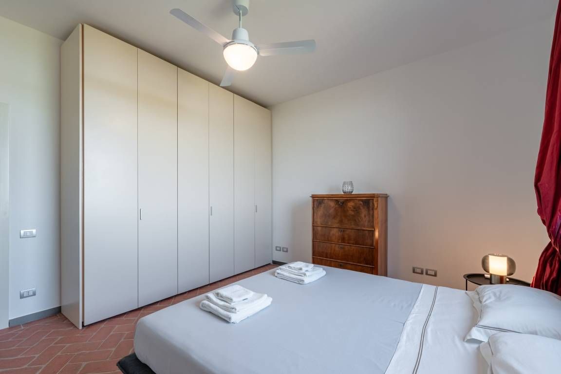 A hotel room with a bed, a wardrobe, a wooden cabinet, a ceiling fan, and a nightstand with a lamp.