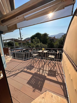Um deck de madeira com guarda-corpos de metal, piscina e vista para uma área residencial com casas e colinas ao fundo.