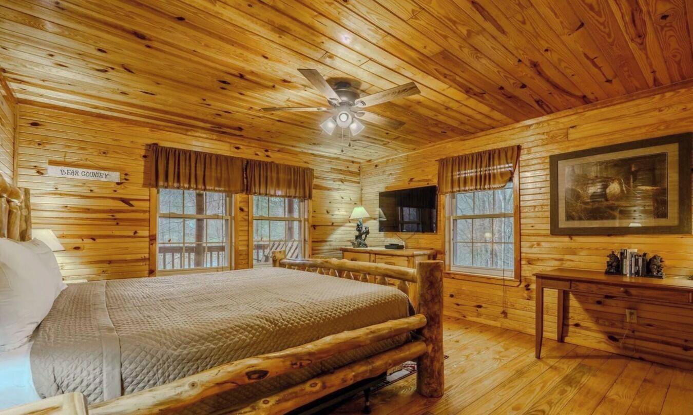 A log cabin bedroom with a bed, a ceiling fan, a painting, and a window with curtains.