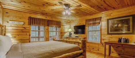 A log cabin bedroom with a bed, a ceiling fan, a painting, and a window with curtains.