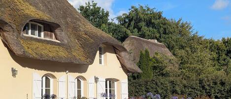 A traditional house with a thatched roof and white shutters, surrounded by a garden with various plants and flowers.