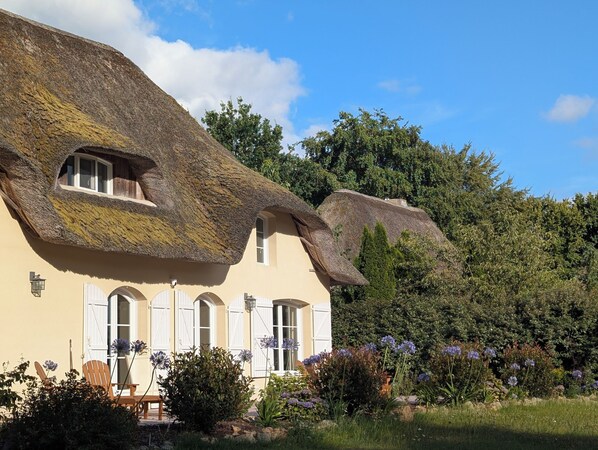 A traditional house with a thatched roof and white shutters, surrounded by a garden with various plants and flowers.