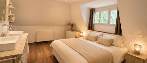 Un dormitorio con una cama grande, mesitas de noche, una ventana con cortinas y un baño con lavamanos y espejo.