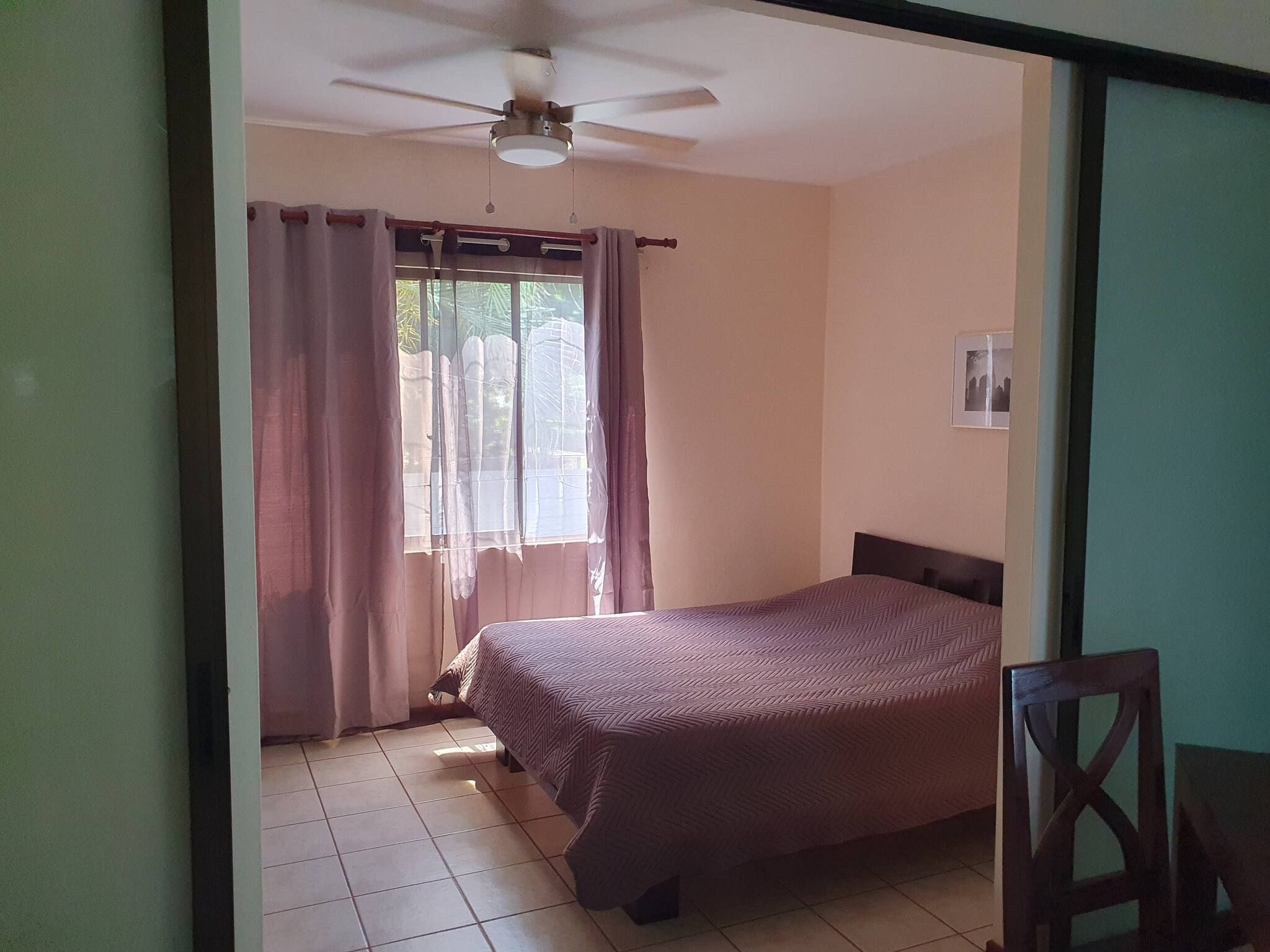 Quarto com cama, ventilador de teto, janela com cortinas e piso ladrilhado.
