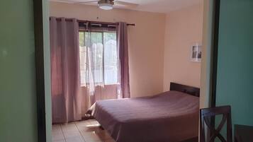 Quarto com cama, ventilador de teto, janela com cortinas e piso ladrilhado.