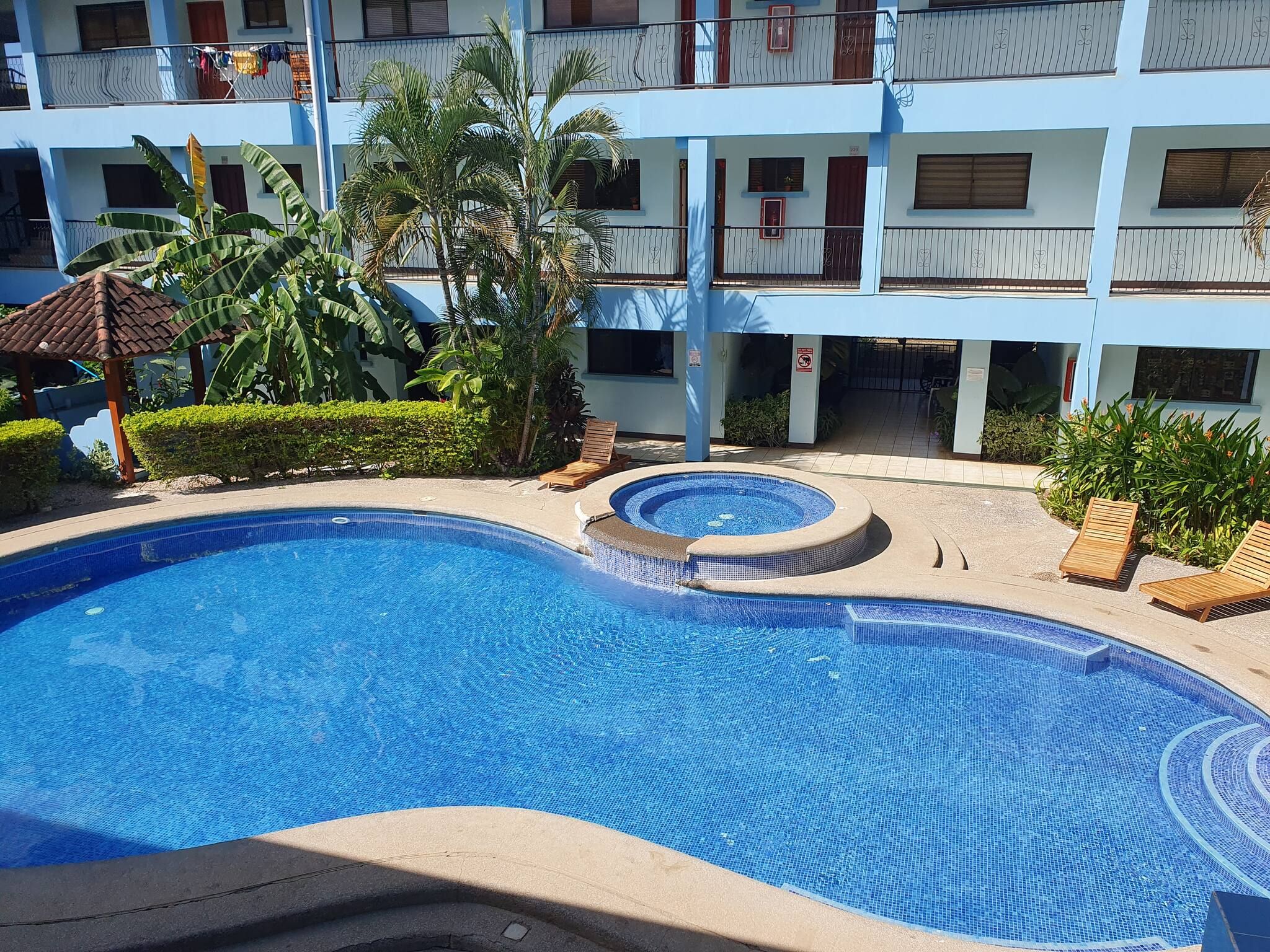 A swimming pool with a hot tub and lounge chairs, surrounded by a building with multiple balconies and tropical plants.