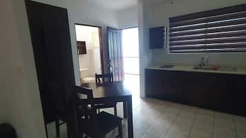 Un comedor con mesa y sillas, un ventilador de techo y una zona de cocina con ventana y persianas.