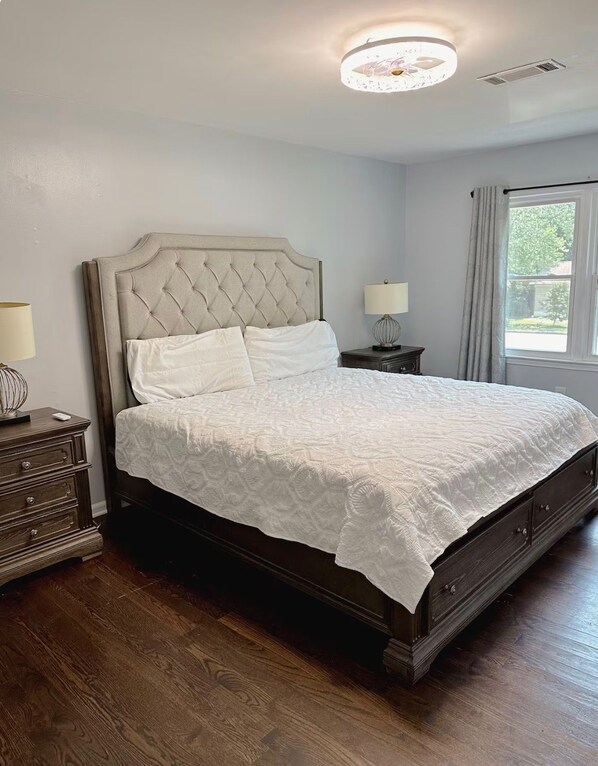 Un dormitorio con una cama grande, una mesita de noche, una lámpara y una ventana con cortinas.