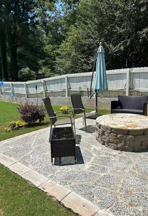 Un área de patio al aire libre con piso de piedra, dos sillones de descanso, una mesa redonda de piedra y un paraguas azul.