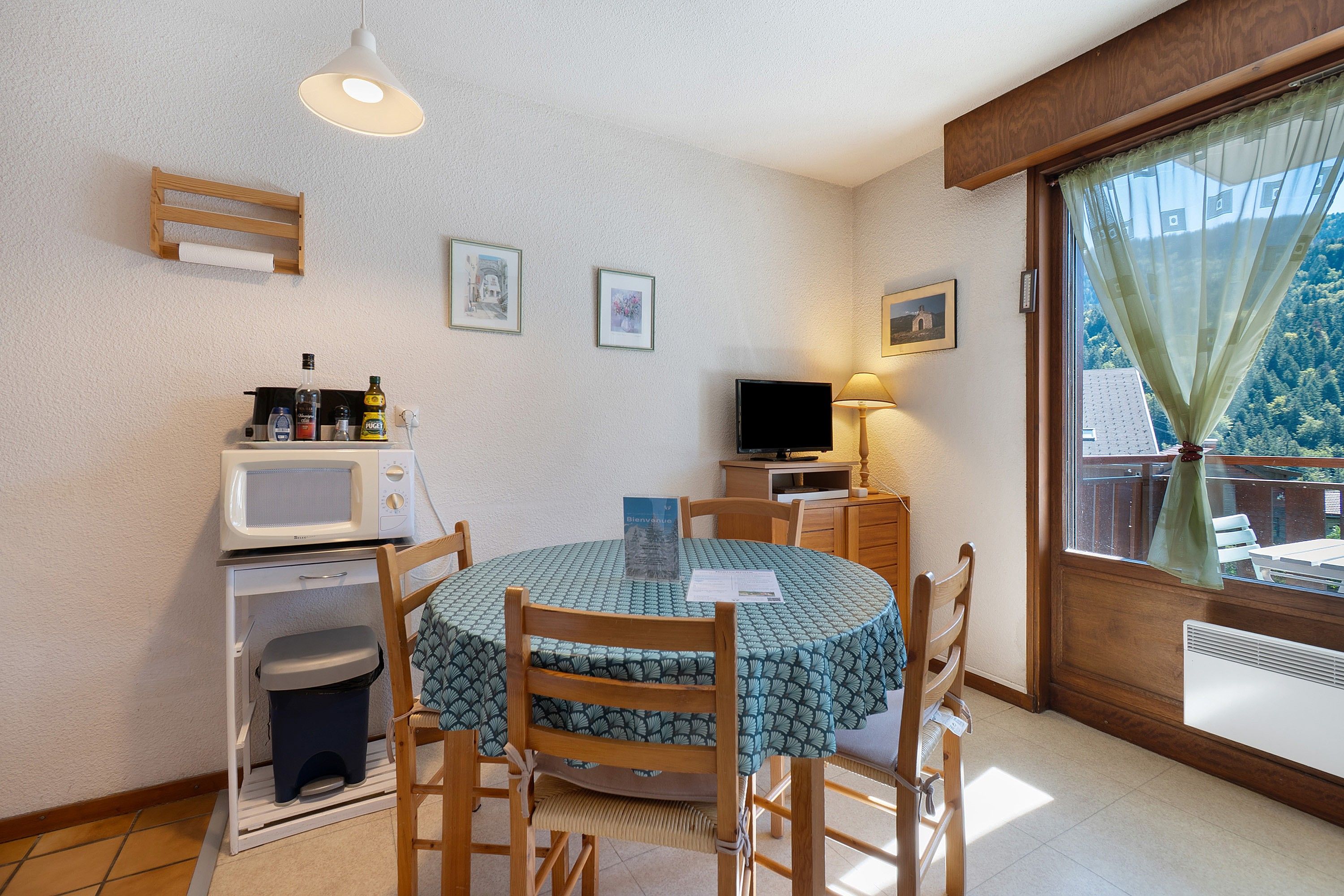 Une cuisine compacte équipée d’un four à micro-ondes, d’un évier et d’un petit réfrigérateur. On y trouve une table à manger avec des chaises, un téléviseur et une fenêtre avec un rideau.