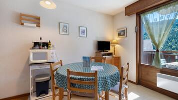 Une cuisine compacte équipée d’un four à micro-ondes, d’un évier et d’un petit réfrigérateur. On y trouve une table à manger avec des chaises, un téléviseur et une fenêtre avec un rideau.