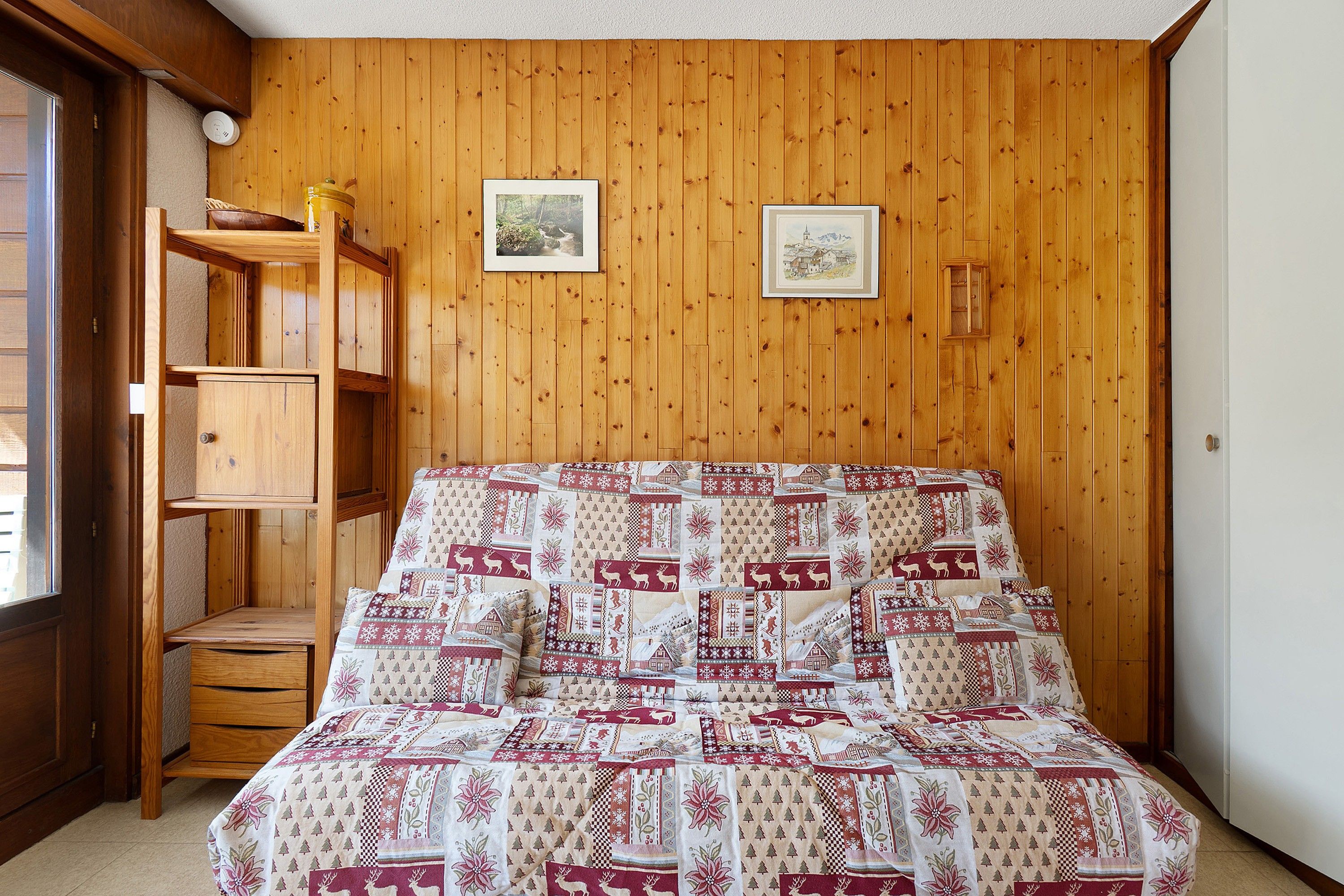 A bedroom with a wooden paneled wall, a bed with a patterned quilt, a wooden shelf unit, and two framed pictures.