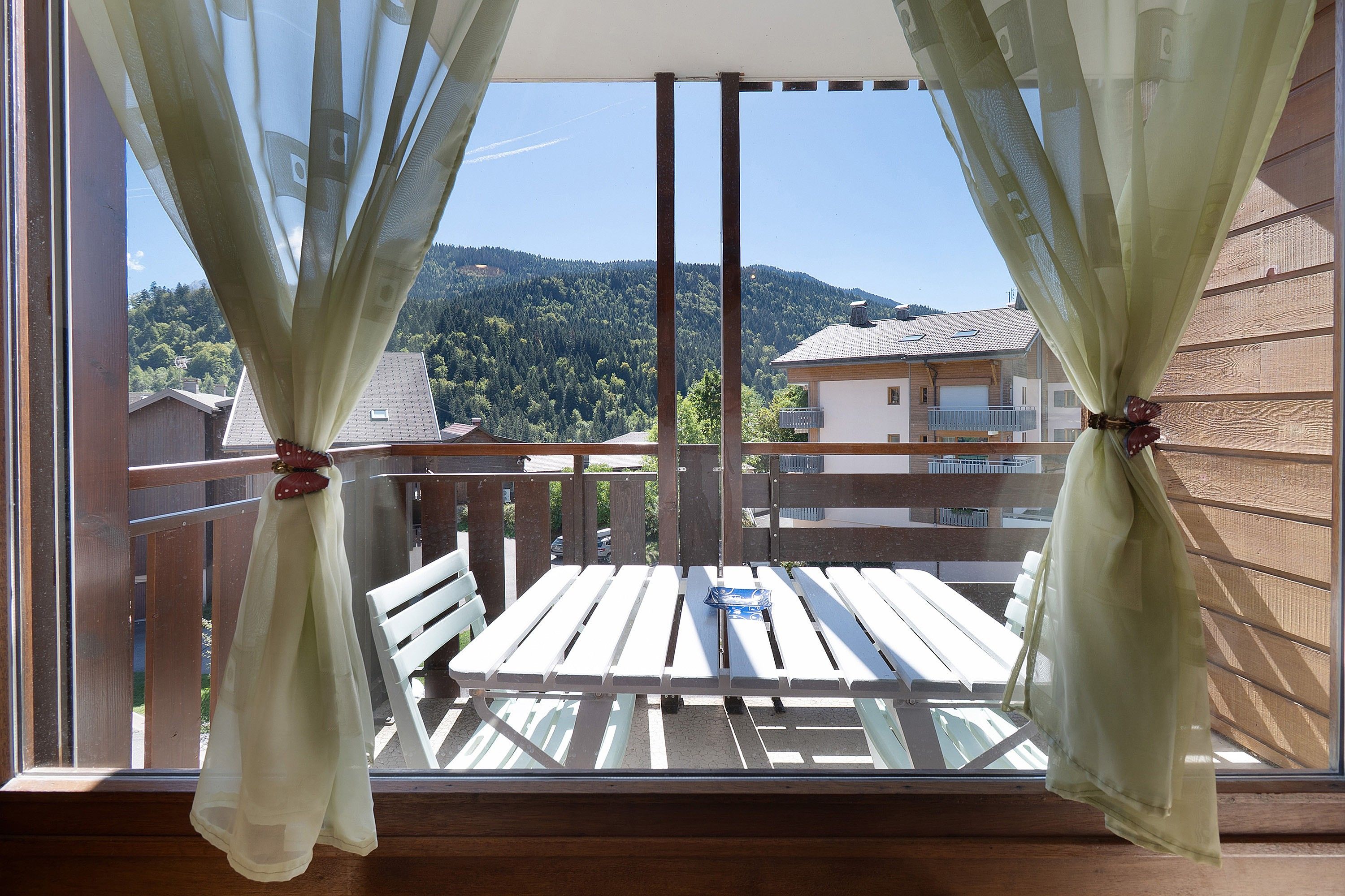 A balcony with a wooden table and chairs, curtains tied with a bow, and a view of a forested hillside.