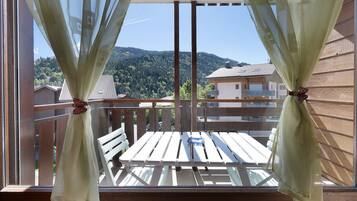 A balcony with a wooden table and chairs, curtains tied with a bow, and a view of a forested hillside.