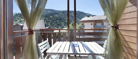 Ein Balkon mit Holztisch und Stühlen, Vorhängen, die mit einer Schleife zusammengebunden sind, und Blick auf einen bewaldeten Hügel.
