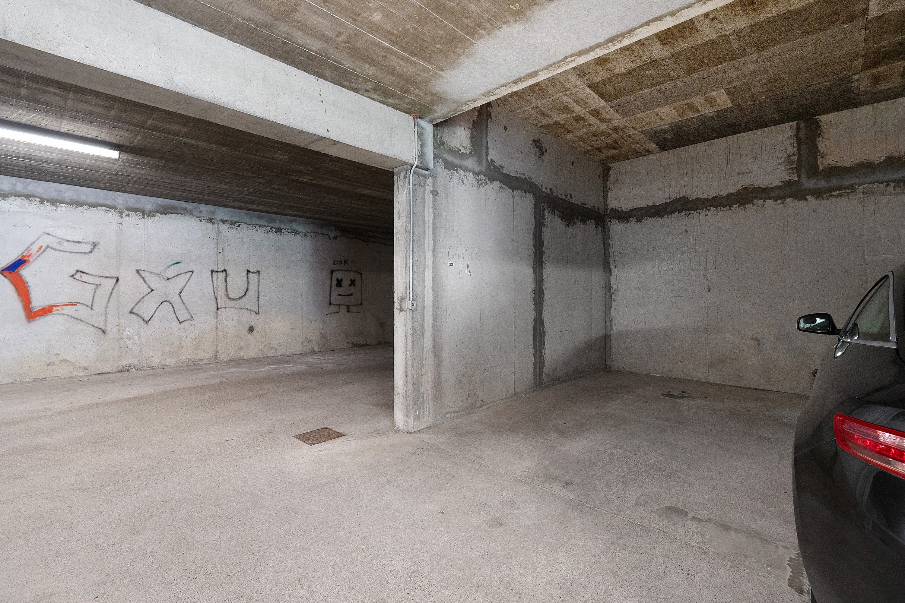 Un parking avec des murs en béton, une voiture garée à droite et des graffitis sur le mur de gauche.