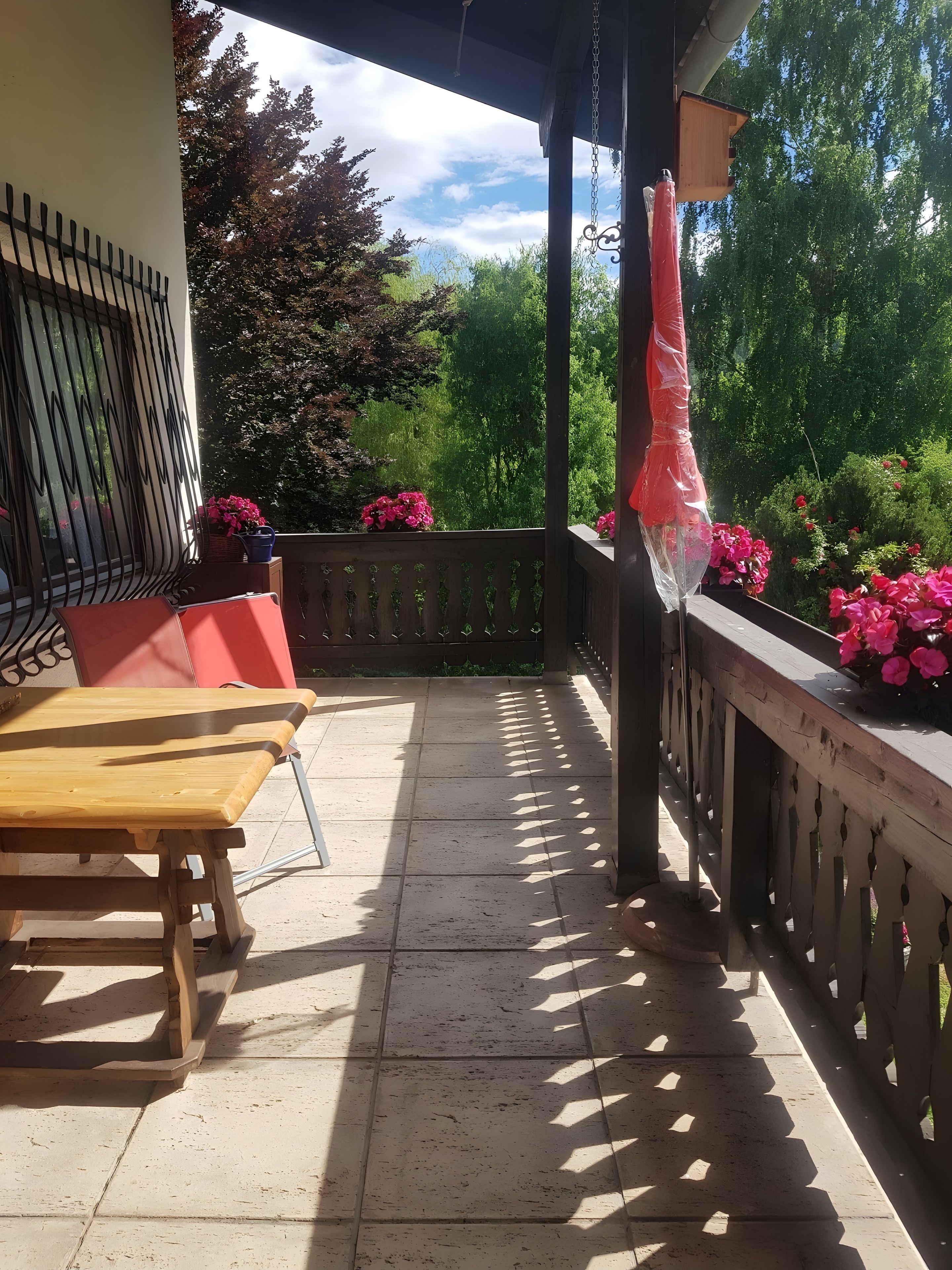 Een overdekt buitengebied met een houten bank, een tafel en een hek dat uitkijkt op een tuin vol roze bloemen.