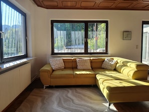 A yellow sectional sofa with patterned cushions, a radiator, a large window with a view of greenery, and a ceiling with wooden paneling.