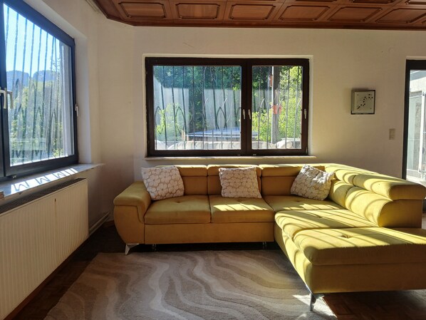 Ein gelbes, modulares Sofa mit gemusterten Kissen, ein Heizkörper, ein großes Fenster mit Blick auf Grünflächen und eine Decke mit Holzvertäfelung.