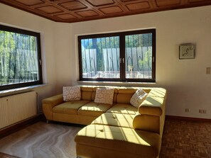 Una habitación con un sofá amarillo, dos ventanas, un radiador y suelo de madera.