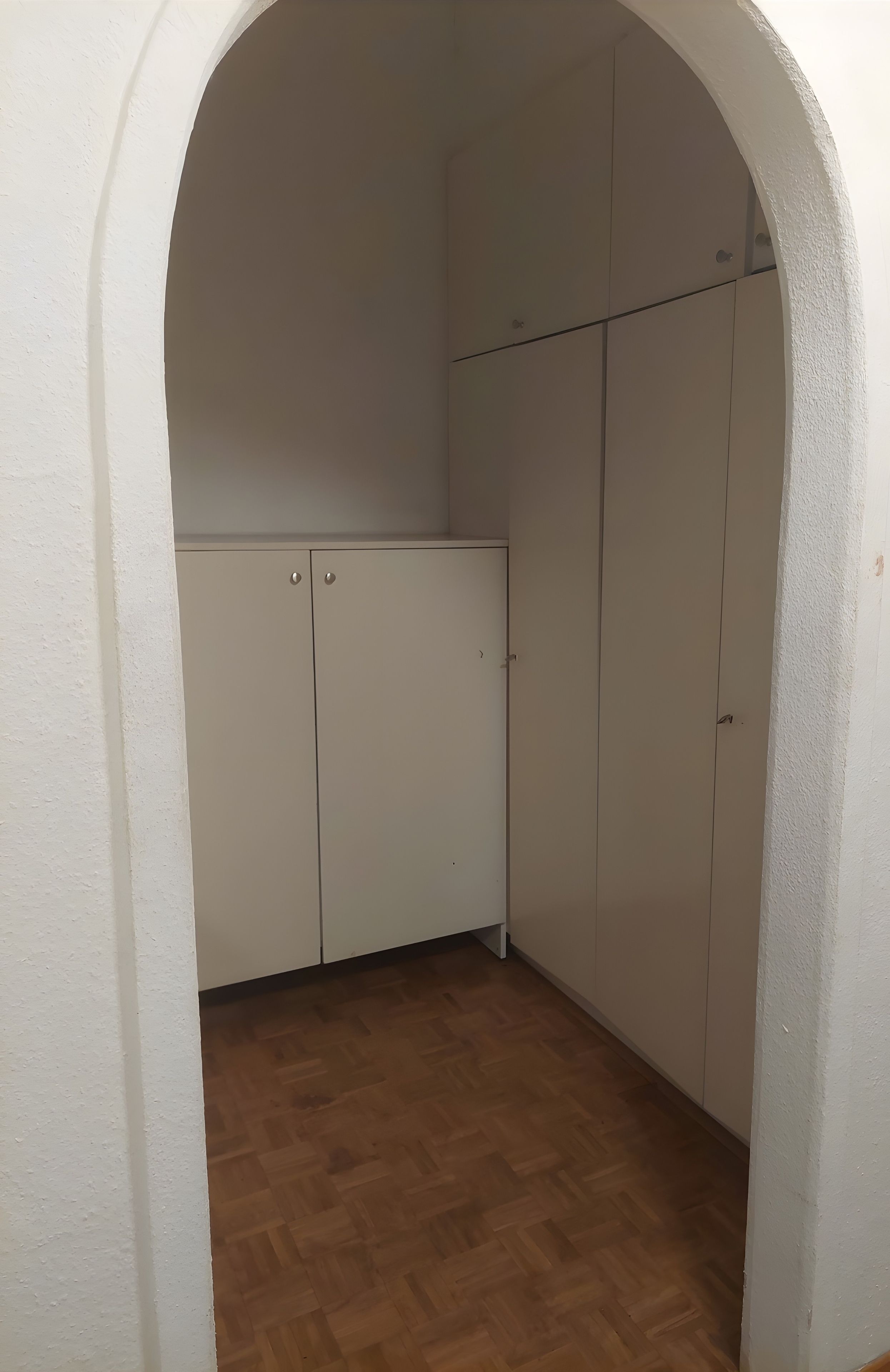 Une pièce dotée d’un plancher en bois, de murs blancs et d’une porte en arc blanc. On y trouve des armoires blanches avec des poignées argentées et un plafond blanc.