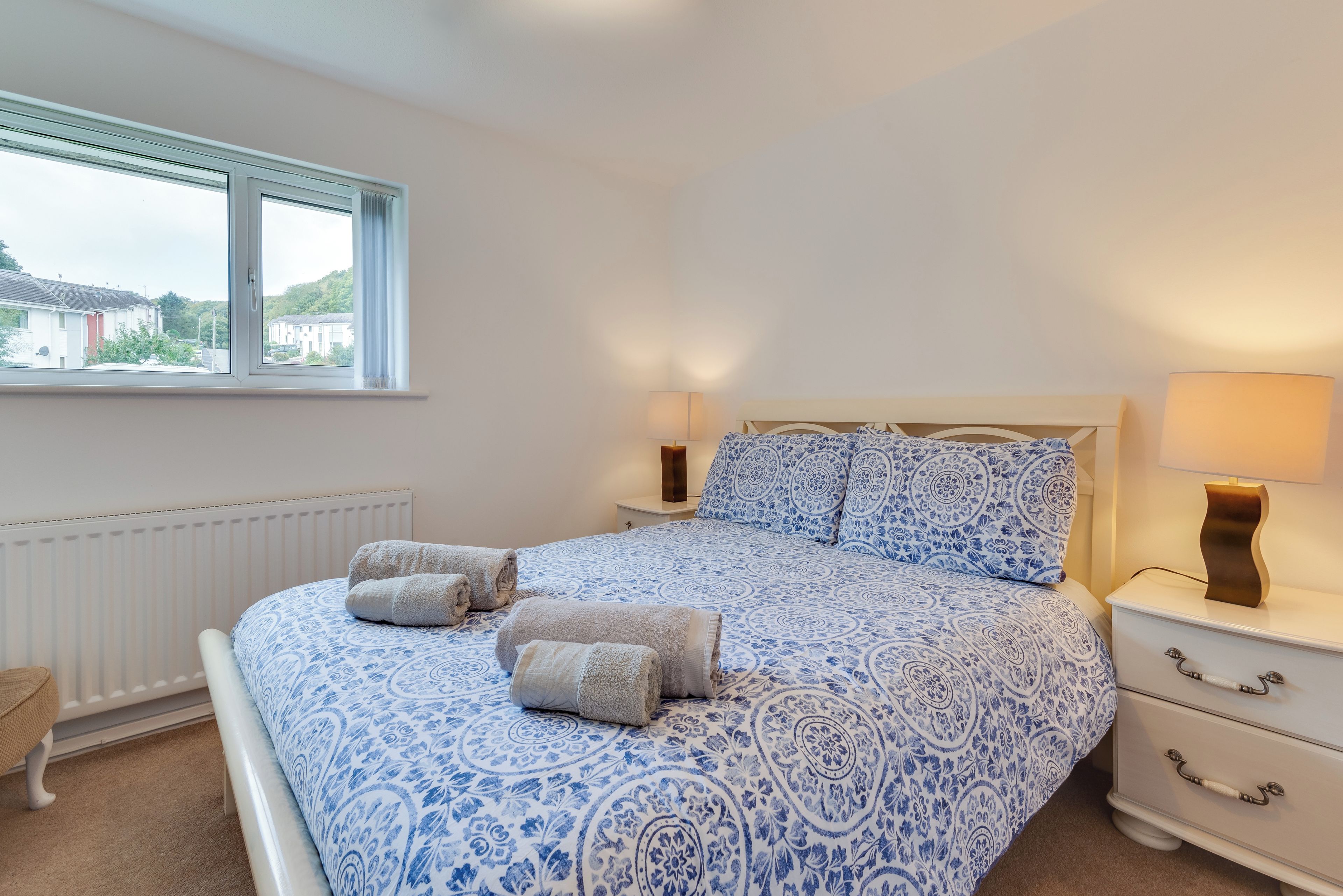 A bedroom with a bed, two bedside lamps, a window, and a radiator.